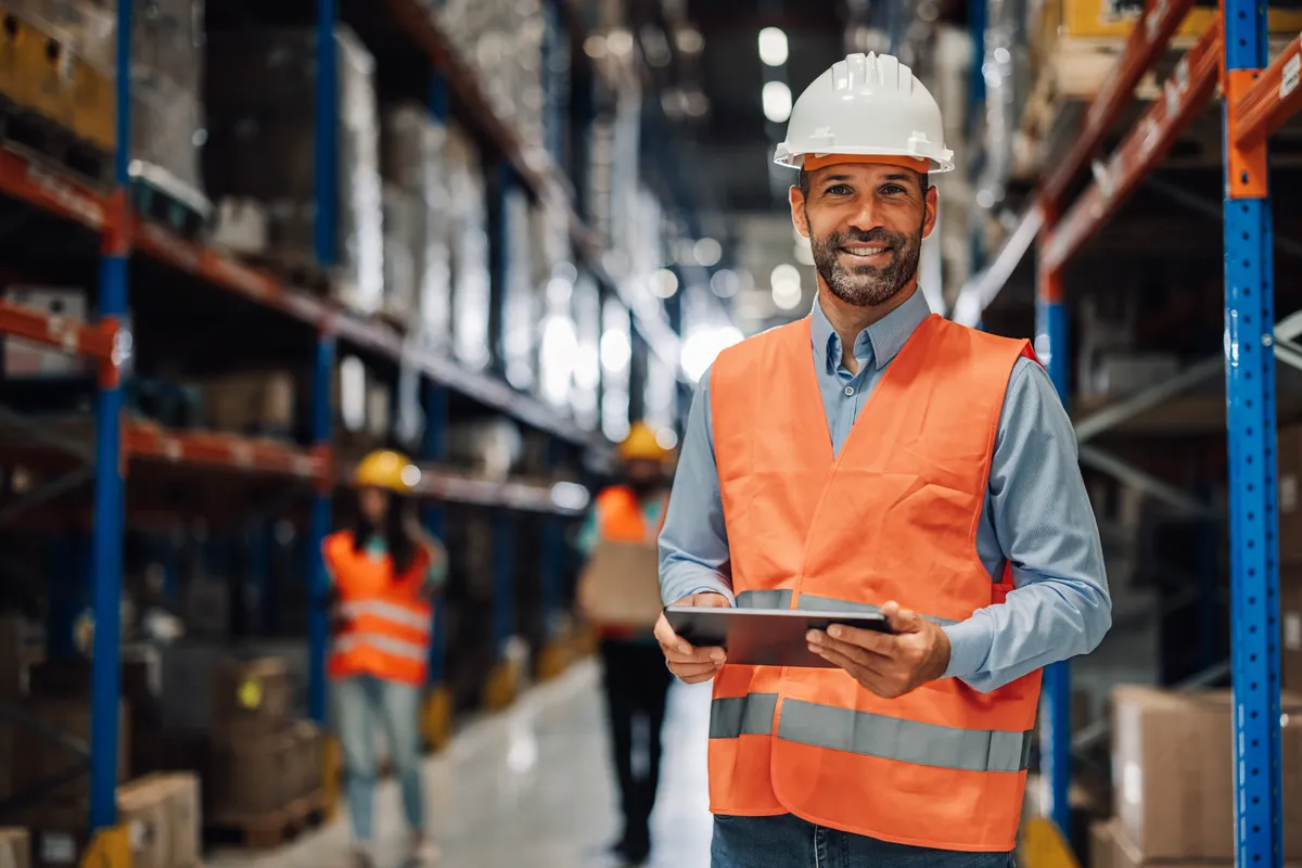 Smiling warehouse worker in a hard hat and hi-vis vest holding a tablet